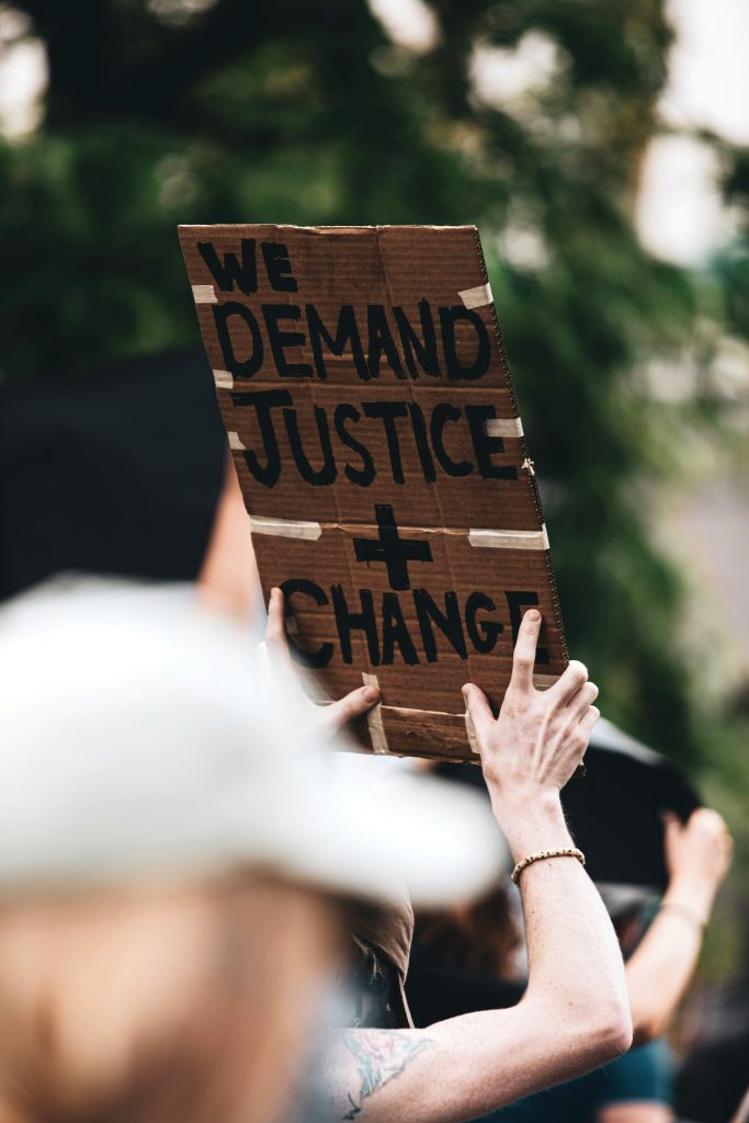 A person holding up a cardboard protest sign that says "We demand justice + change"